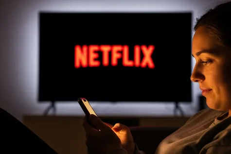 Mujer mirando el móvil con un televisor en el que se ve el logo de Netflix